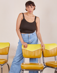 Tiara is standing behind a yellow chair with her hands resting on the back. She is wearing Cropped Cami in Espresso Brown and light wash Sailor Jeans