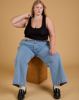 Juliet is sitting on a wooden box wearing Mini Tank in Basic Black and light wash Indigo Wide Leg Trousers