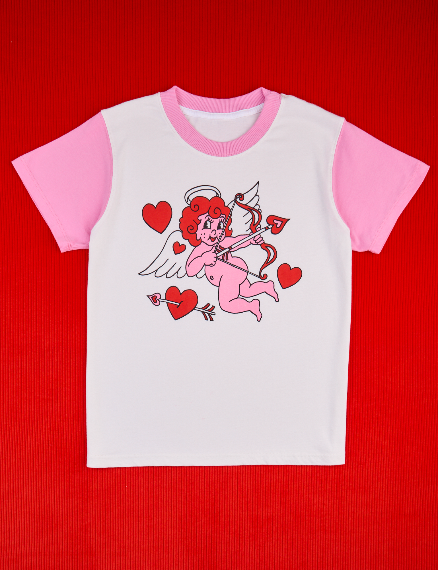 Vintage Tee in Cupid. White body with bubblegum pink sleeves and neckline. Illustrated pink cupid with red hair holding bow and arrow. Red hearts surround cupid.