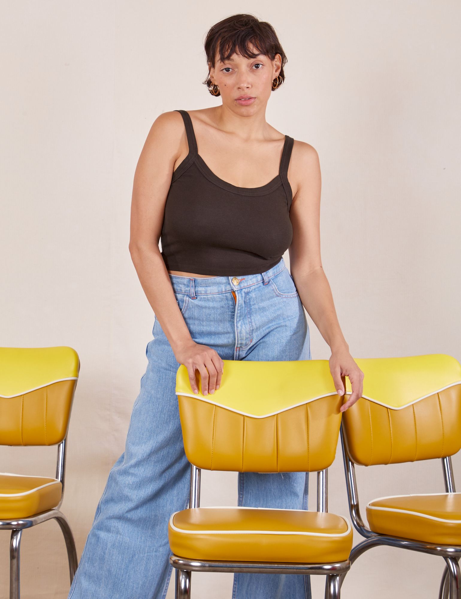Tiara is standing behind a yellow chair with her hands resting on the back. She is wearing Cropped Cami in Espresso Brown and light wash Sailor Jeans