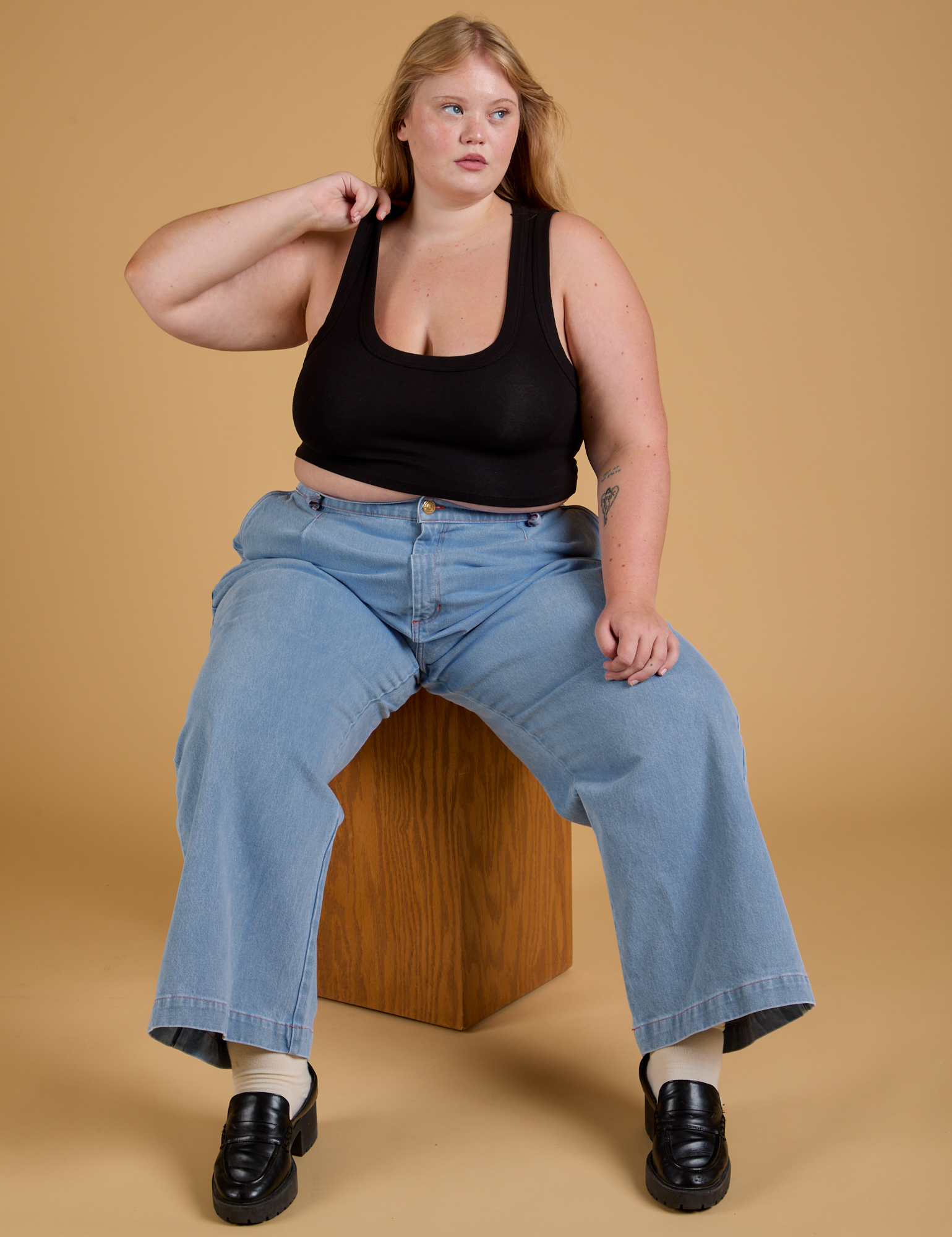 Juliet is sitting on a wooden box wearing Mini Tank in Basic Black and light wash Indigo Wide Leg Trousers