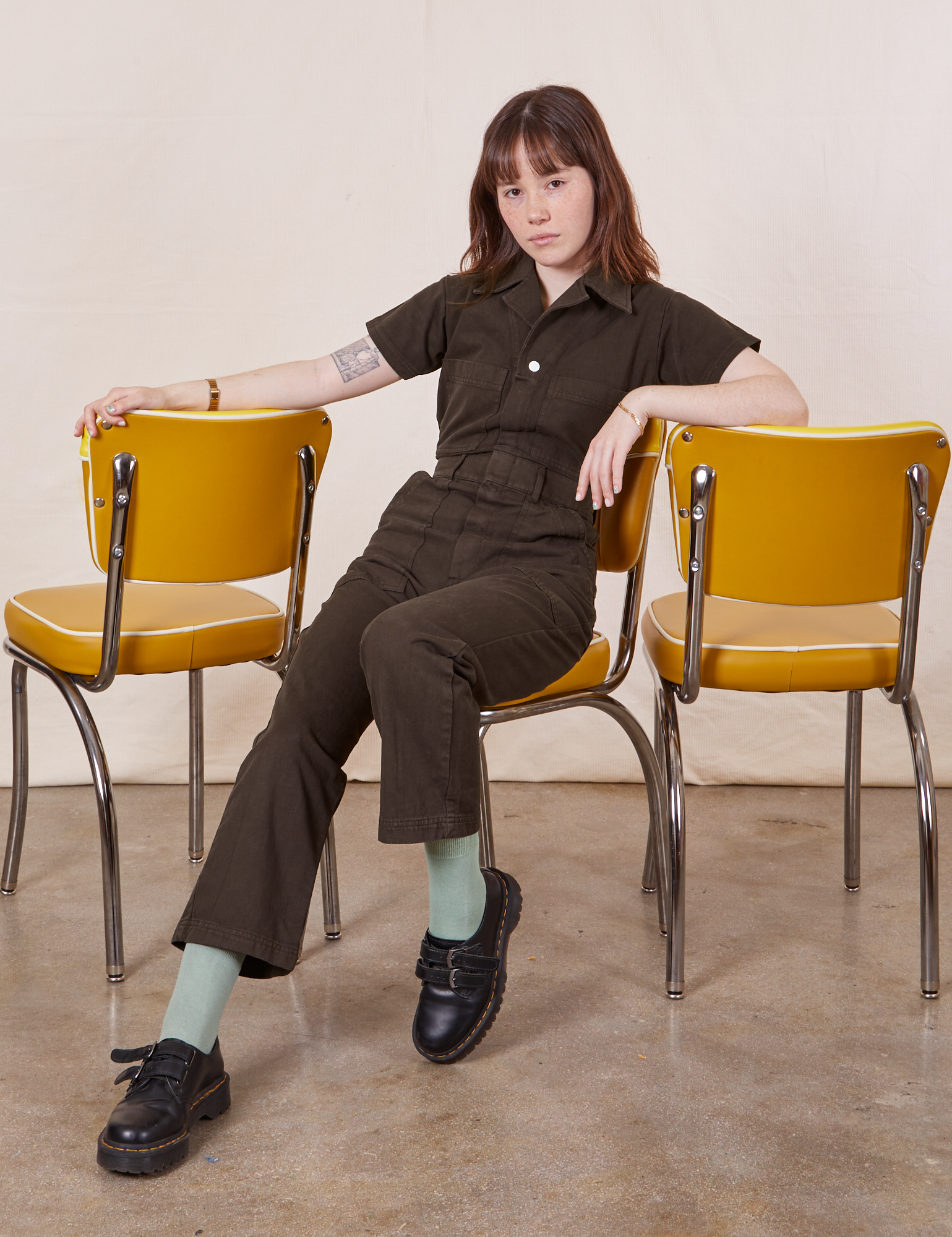 Hana is sitting on a yellow chair wearing Petite Short Sleeve Jumpsuit in Espresso Brown